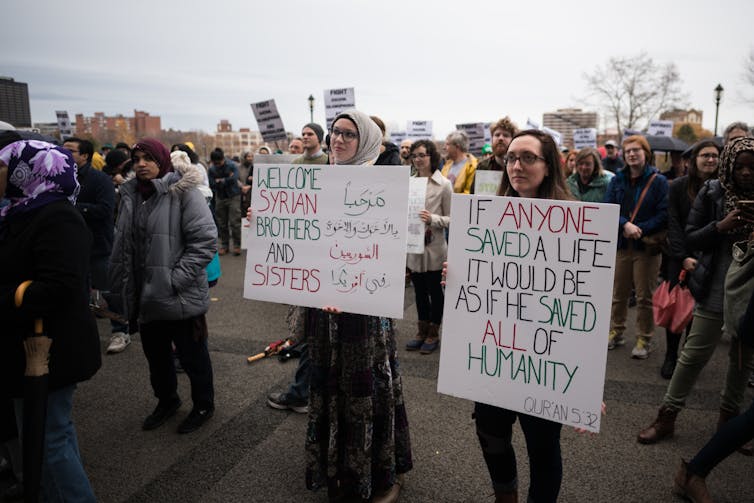 Two women stand on a street holding large signs while people stand behind them.