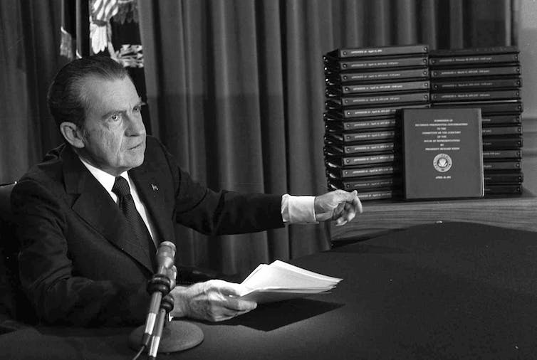 A black and white photo of a man in a suit pointing at a table stacked with bound volumes.