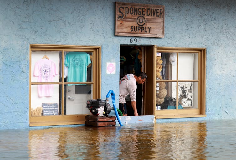 A man looks out a door that is blocked at the bottom. A sump pump is running next to it. The water is nearly up to the windows.