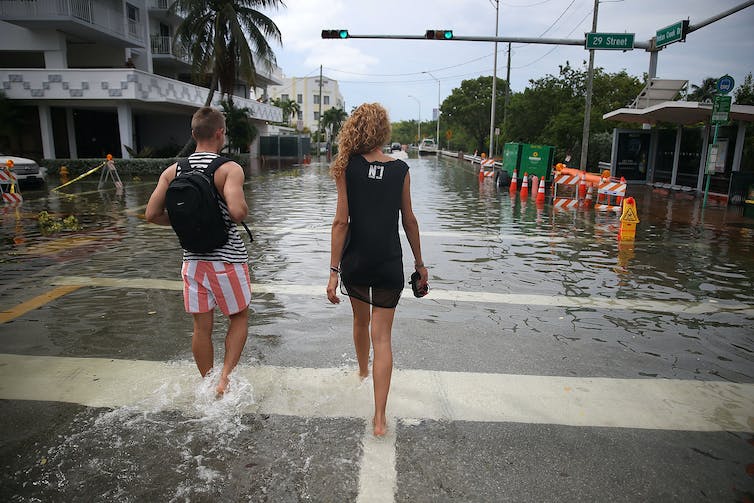 A woman in a dress carrying her shoes and a man in red and white striped shorts walk down a street that is filled with water to above their ankles.