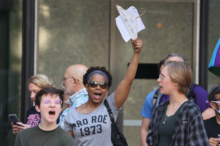 With people standing around her, a woman wearing a shirt that reads, 