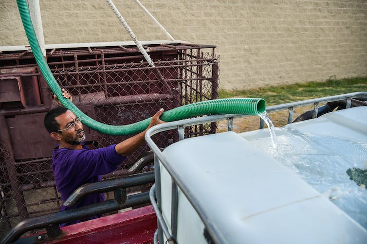 A man squints into the sun as he holds a large hose that pours water into a tank in the back of a pickup truck.