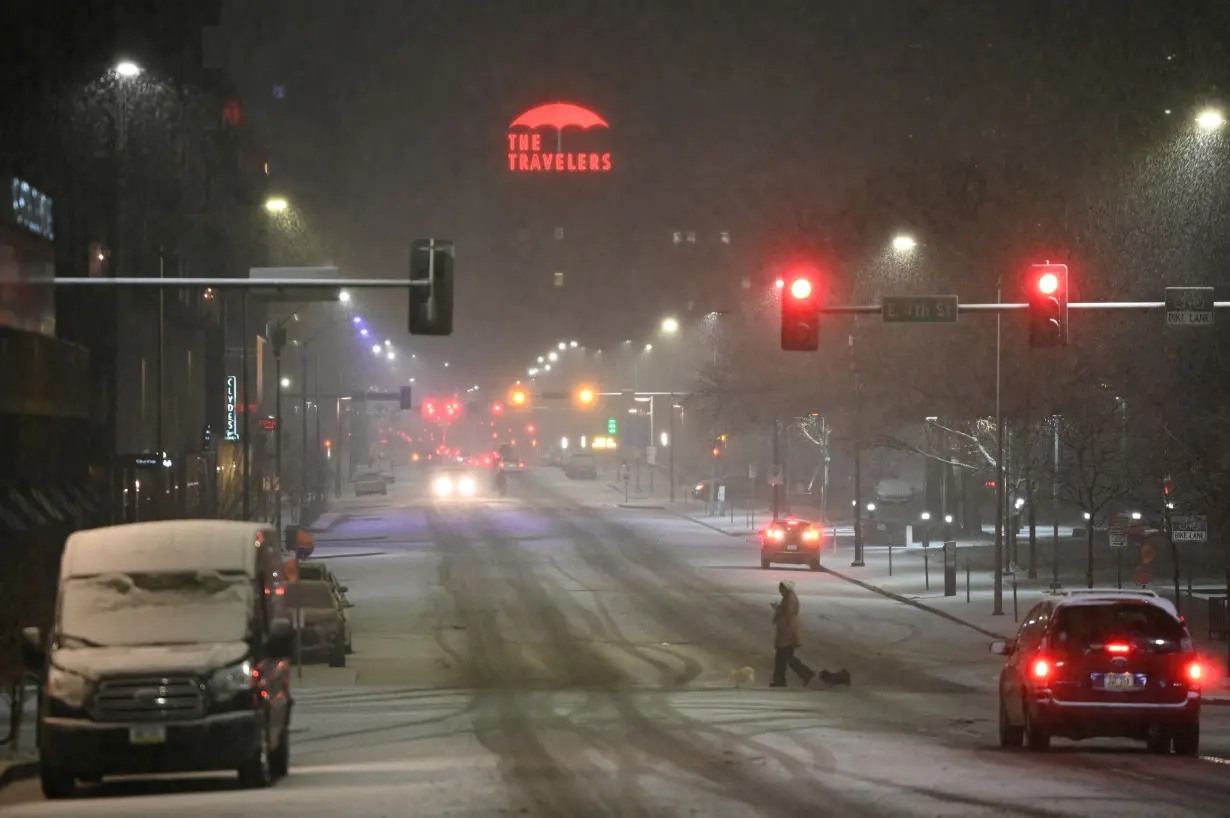 A person walks dogs across a road while cars drive by as it snows in Des Moines