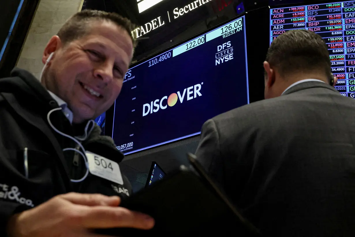 A screen displays the logo and trading information for Discover Financial as traders work on the floor at the NYSE in New York