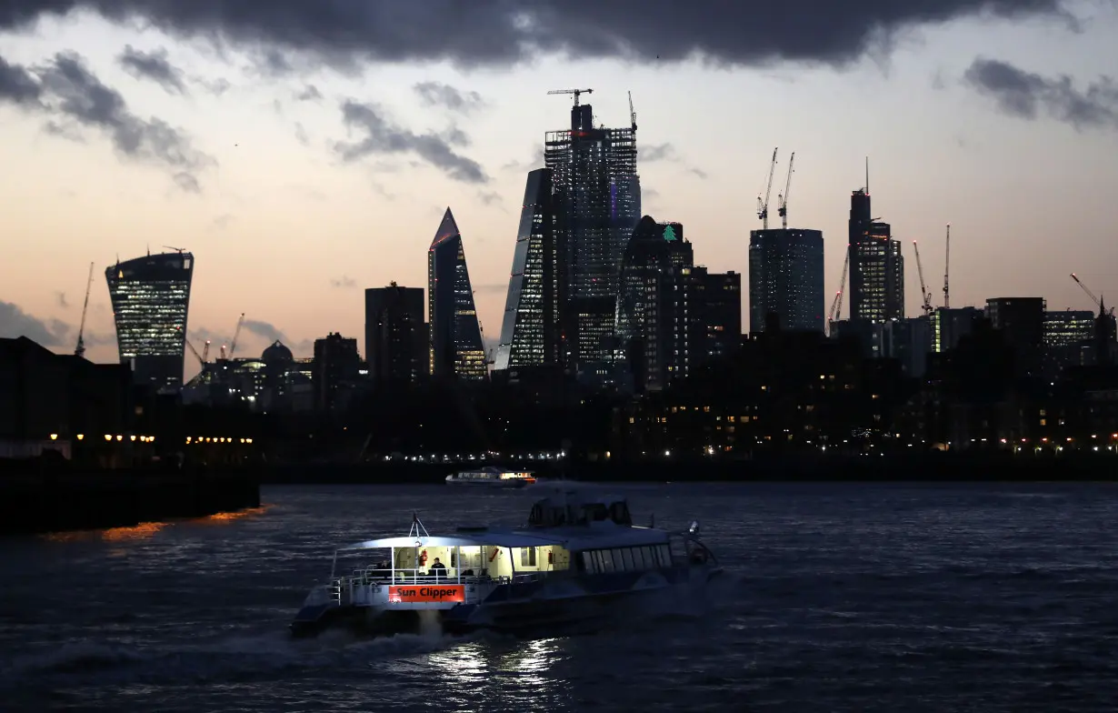 A river boat cruises down the River Thames as the sun sets behind the Canary Wharf financial district of London