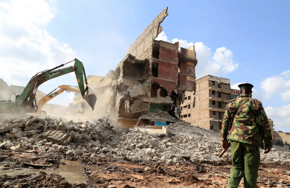 A riot police officer stands guard as an excavator clears the rubble during the search and rescue operations of a residential flat built on riparian land, that collapsed while undergoing demolition in Nairobi