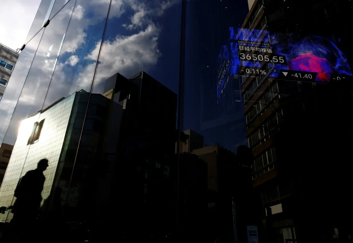 A pedestrian is reflected on a glass of a business building while an electric board showing Nikkei index is seen in the building at a business district in Tokyo