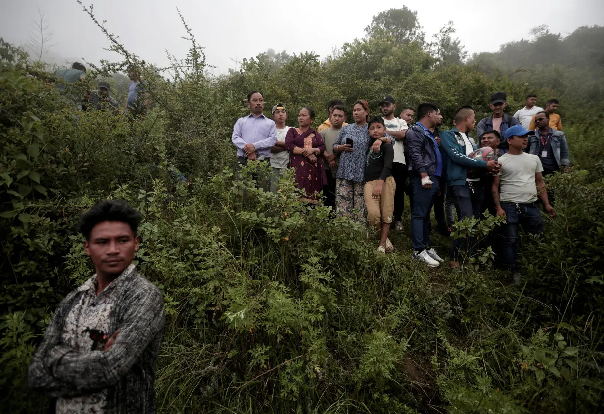 People watch as security force personnel prepare to carry the bodies of the victims at the site of a helicopter crash on the outskirts of Kathmandu