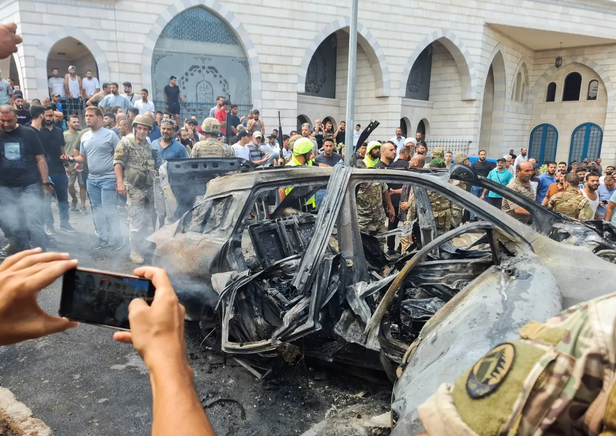 A man uses his phone as he stands near a burnt car after an Israeli strike on the outskirts of the southern port city of Sidon, according to two Palestinian sources