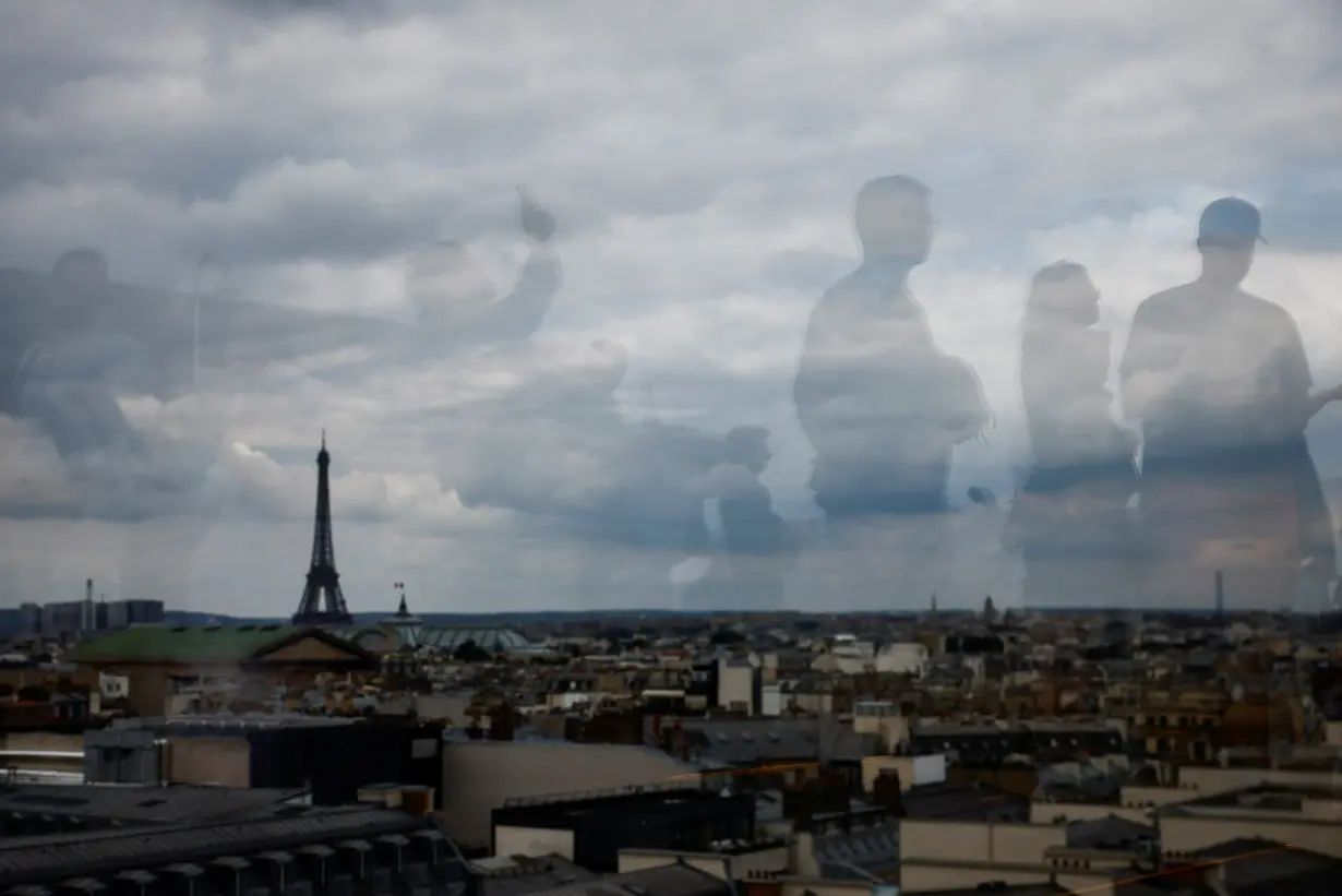 Tourists are reflected on the balustrade as they look at the Eiffel Tower and the Paris skyline from the rooftop of the Galeries Lafayette department store on a cloudy day in Paris