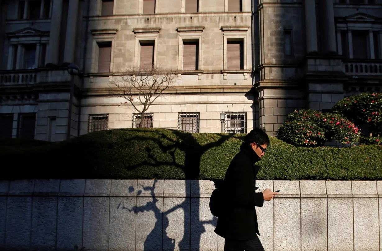 FILE PHOTO: A man using his mobile phone walks past the Bank of Japan headquarters building in Tokyo