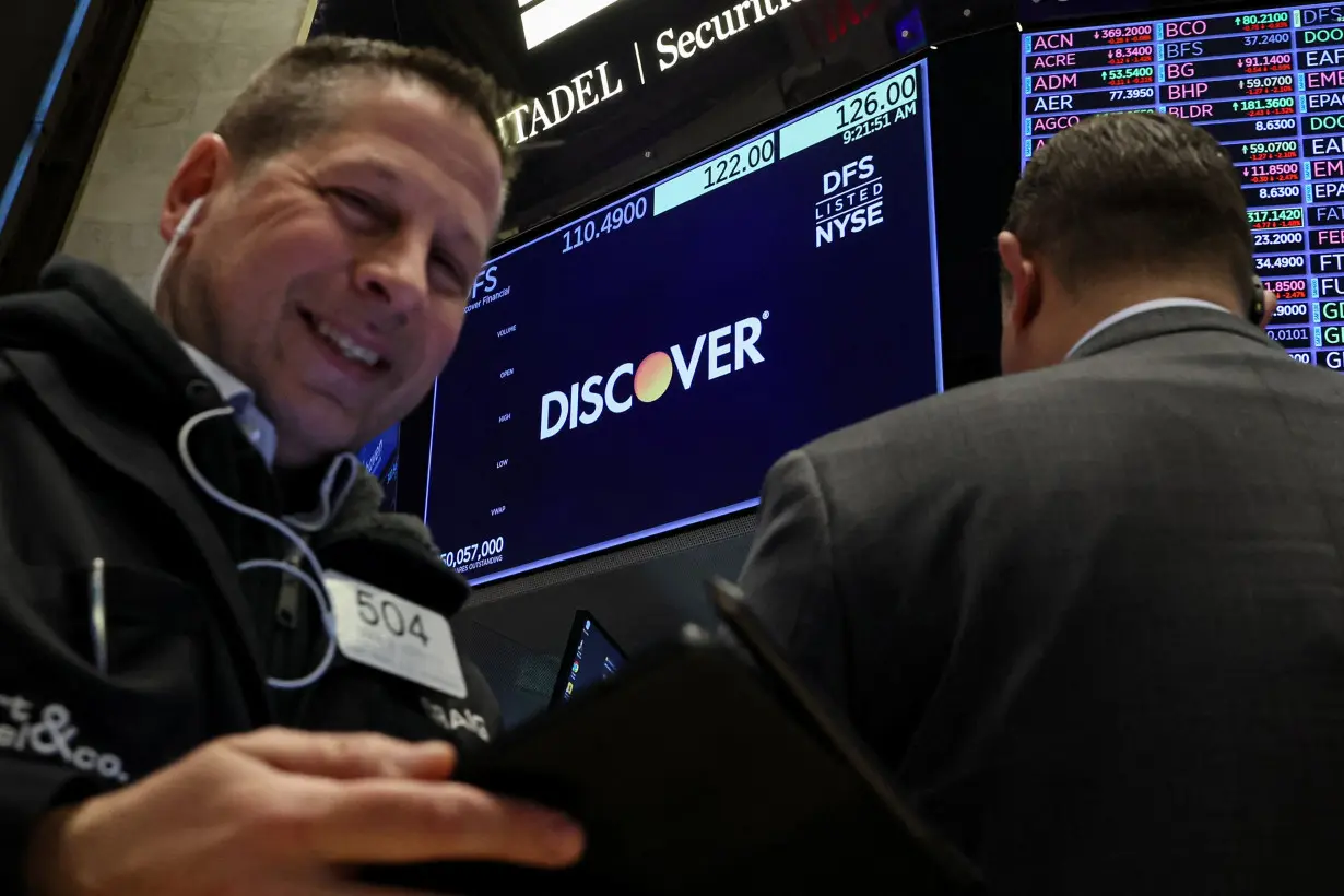 A screen displays the logo and trading information for Discover Financial as traders work on the floor at the NYSE in New York