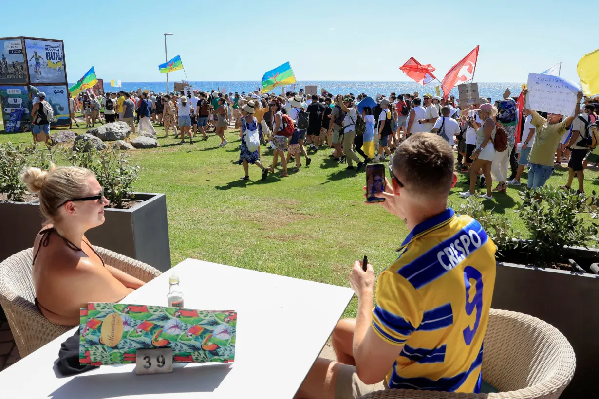 A couple of tourists sitting on a terrace watch a demonstration for a change in the tourism model in the Canary Islands