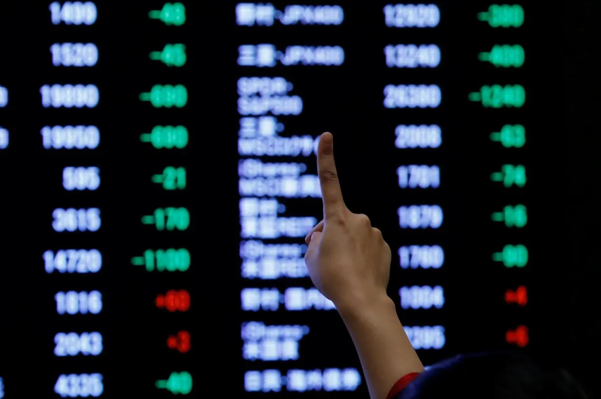 FILE PHOTO: A woman points to an electronic board showing stock prices as she poses in front of the board at the Tokyo Stock Exchange